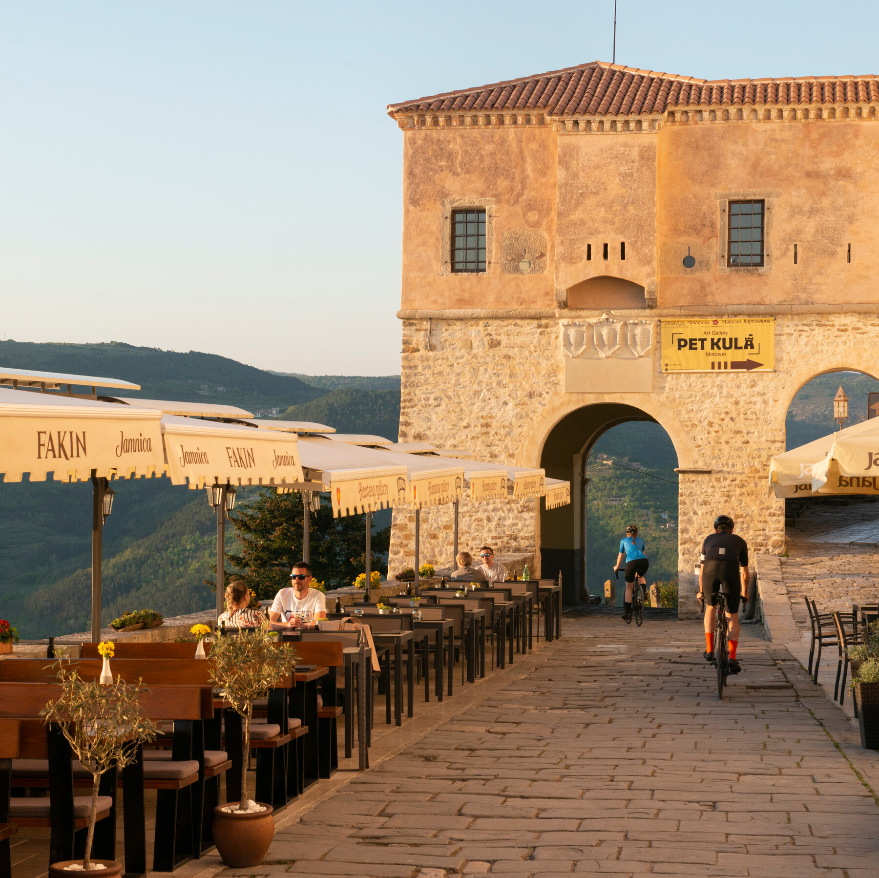 Two people ride their bicycles through a path near a restaurant on top of a mountain.